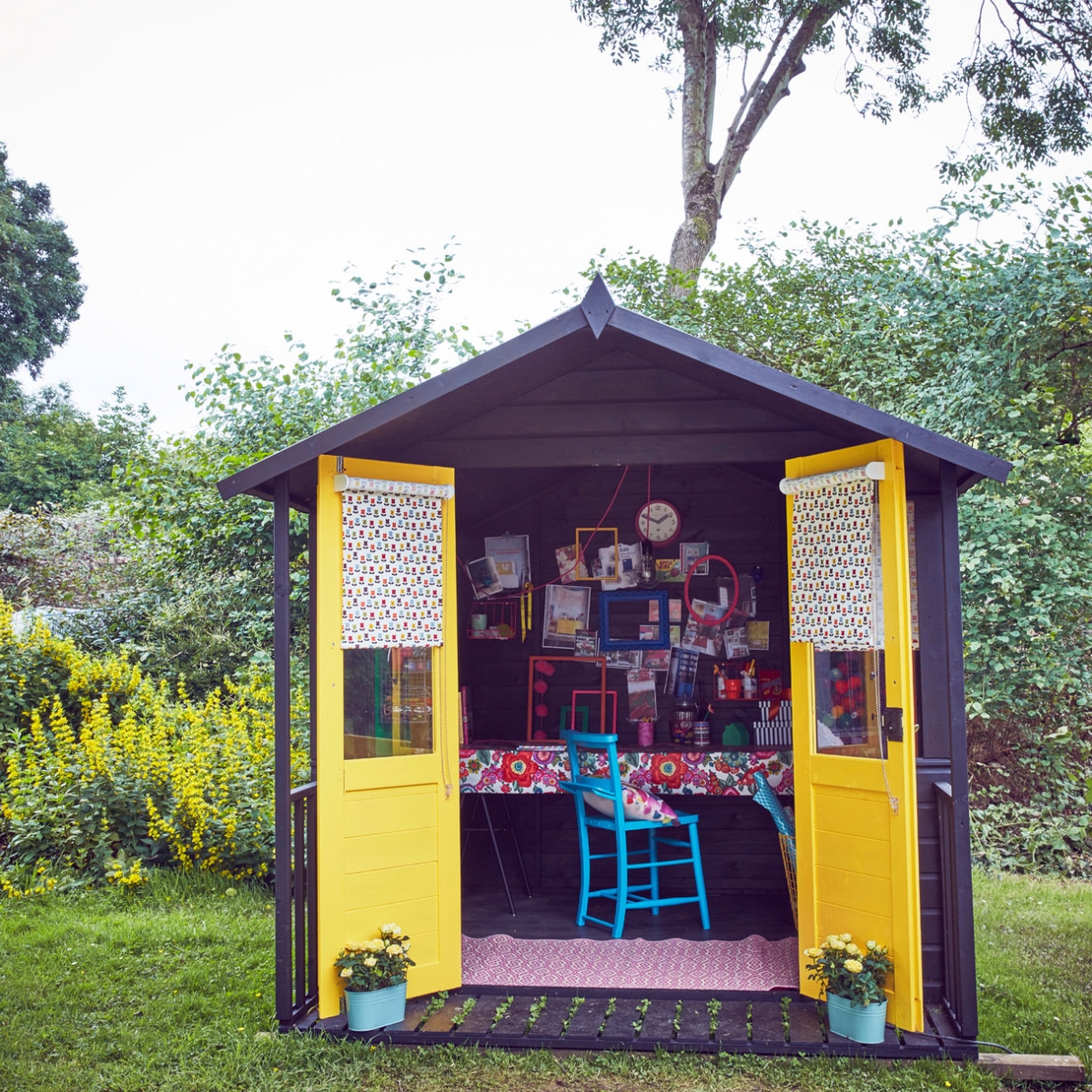 A She shed designed as a crafting DIY and upcycling creaive workspace at the end of the garden. The balck weatherboard exterior is set off by bright yellow doors and patterned fabric blinds