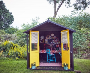 A She shed designed as a crafting DIY and upcycling creaive workspace at the end of the garden. The balck weatherboard exterior is set off by bright yellow doors and patterned fabric blinds