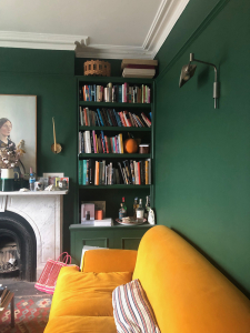 The rich forest green walls and bright yellow sofa add character to the living room, with mini library. Part of Laura Jackson's house tour with Sophie Robinson and Kate Watson-Smyth #thegreatindoors #podcast #sophierobinson #livingroom