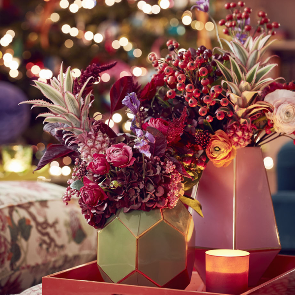 Red tray with green geometric vase and red theme floral display with pineapple tops and berries and Christmas tree in the background. Sophie Robinson's living room