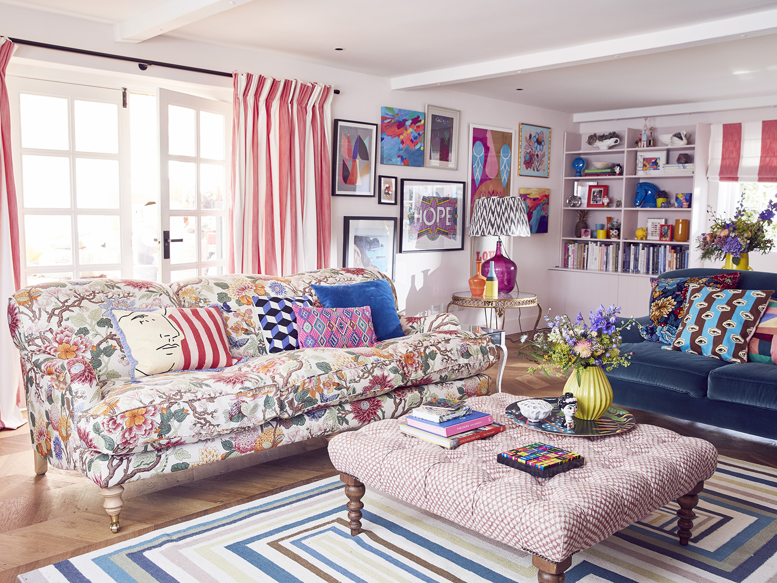 A large bright living room with double doors and red and white striped curtains. A mix of vibrant cushions sit on a floral sofa and dark blue sofa, with a large footstool in front. The walls have a fun gallery wall and bookshelves displaying colourful decorative items. 
