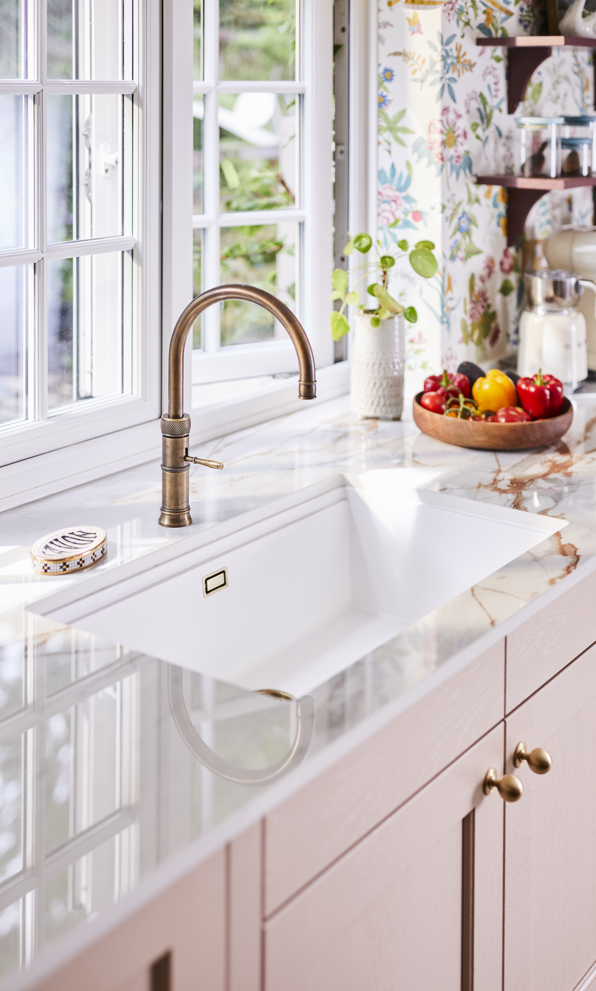 Quooker brass boiling water tap and white sink in front of an open window looking out on the garden. A white worktop with gold vein running through it. 