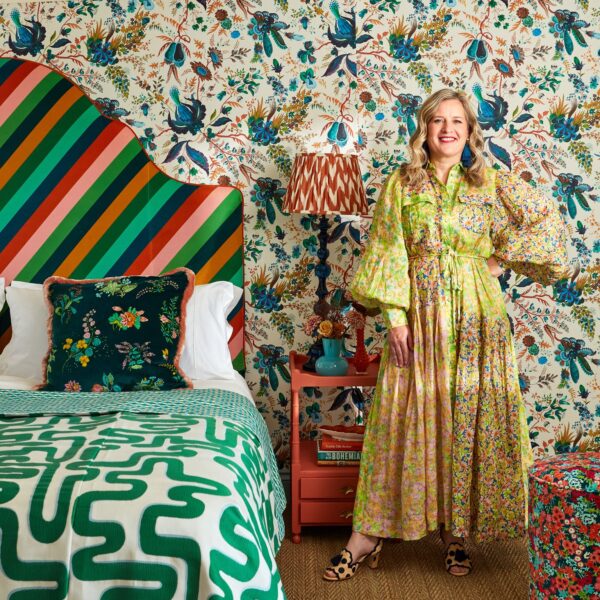 Sophie stands in a gold floral dress next to her bed with wiggle bedspread in emerald green and headboard in diagonal stripe in multiple colours. Behind her is a vintage bedside table with colourful lamp and vase and the white floral wallpaper with blue accents.