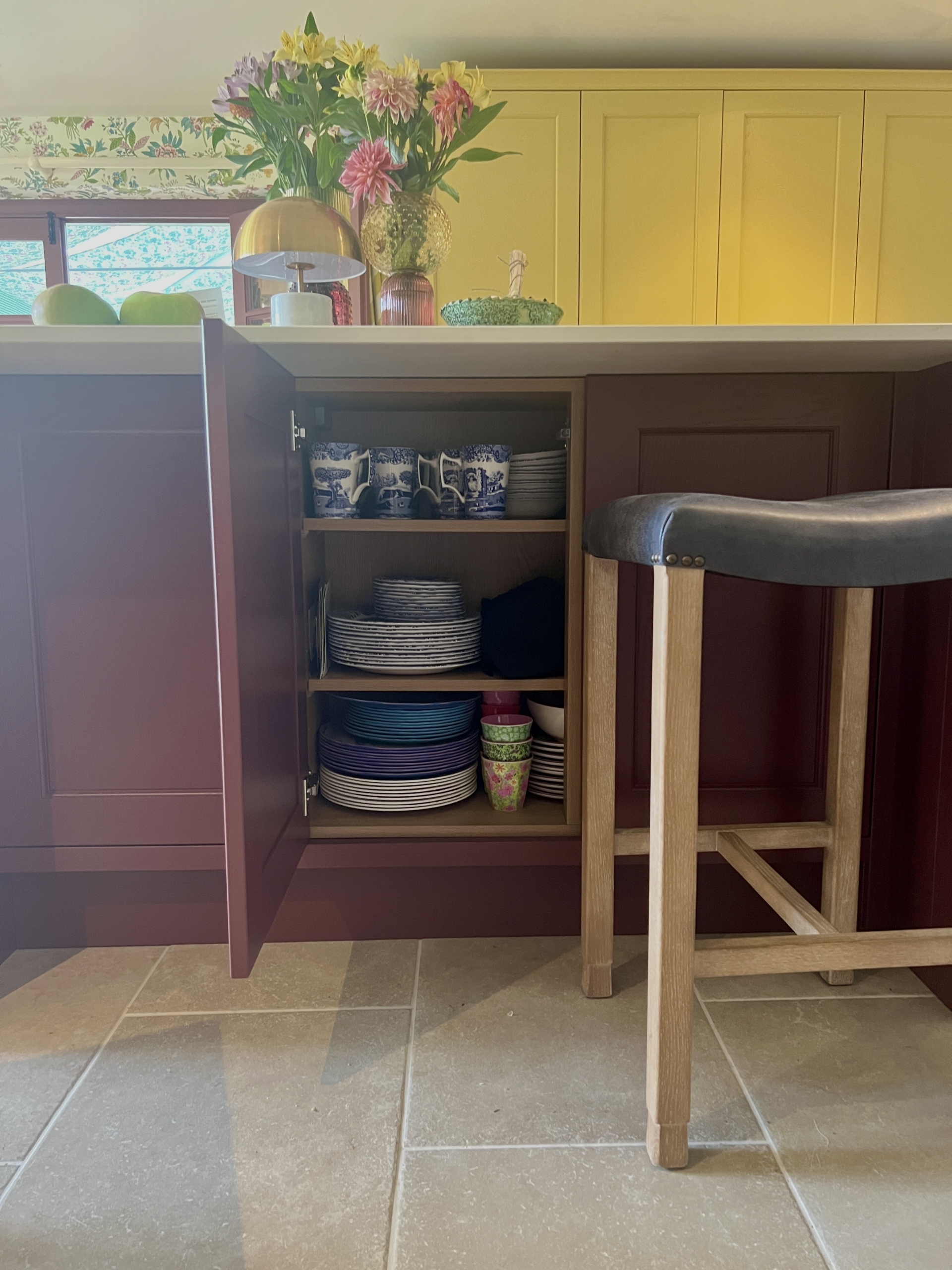Open cupboard door in the kitchen island, displaying plate and mug storage within. Next to the open cupboard is a bar stool with wooden legs and dark leather seat.