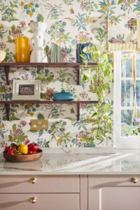 Open shelving on floral wallpaper. Shelves display pictures, vases and plants with a bowl of colourful fruit and veg on the white worktop.
