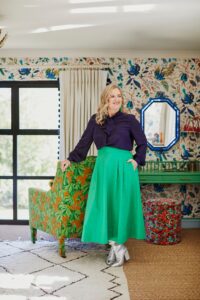 Sophie stands with hand resting on the back of a green armchair. Dressed in a navy blouse and emerald green skirt. The walls have white wallpaper with blue flowers and bird detail, white curtains with blue pom pom trim and a green dressing table with floral fabric stool next to it.