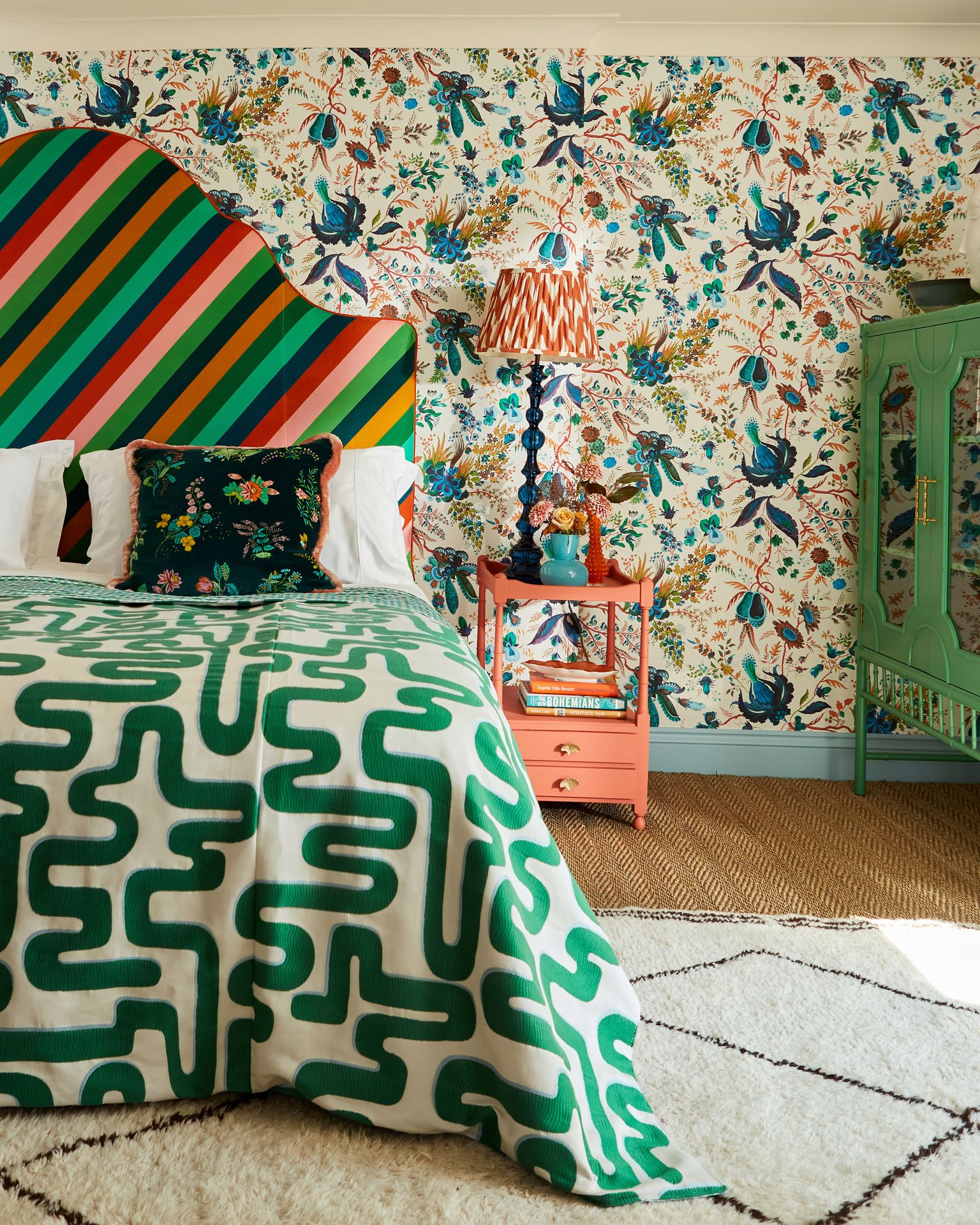 Large double bed with curvy fabric headboard in wide diagonal stripe in rich jewel colours. Bed has white pillows with dark green floral velvet cushion in front of them and a bold bedspread with large green and white squiggle pattern. Rug is soft and white sitting on a sisal style carpet. Bedside table next to the bed is painted coral with tall lamp above it. Walls have white and blue floral wallpaper on them with a blue skirting board. 