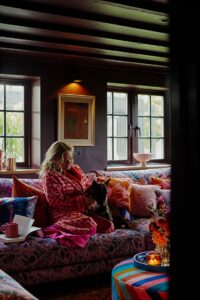 Sophie sits on the sofa in a bath robe, open book and coffee to one side whilst Sophie strokes her dog who is also sat on the sofa.