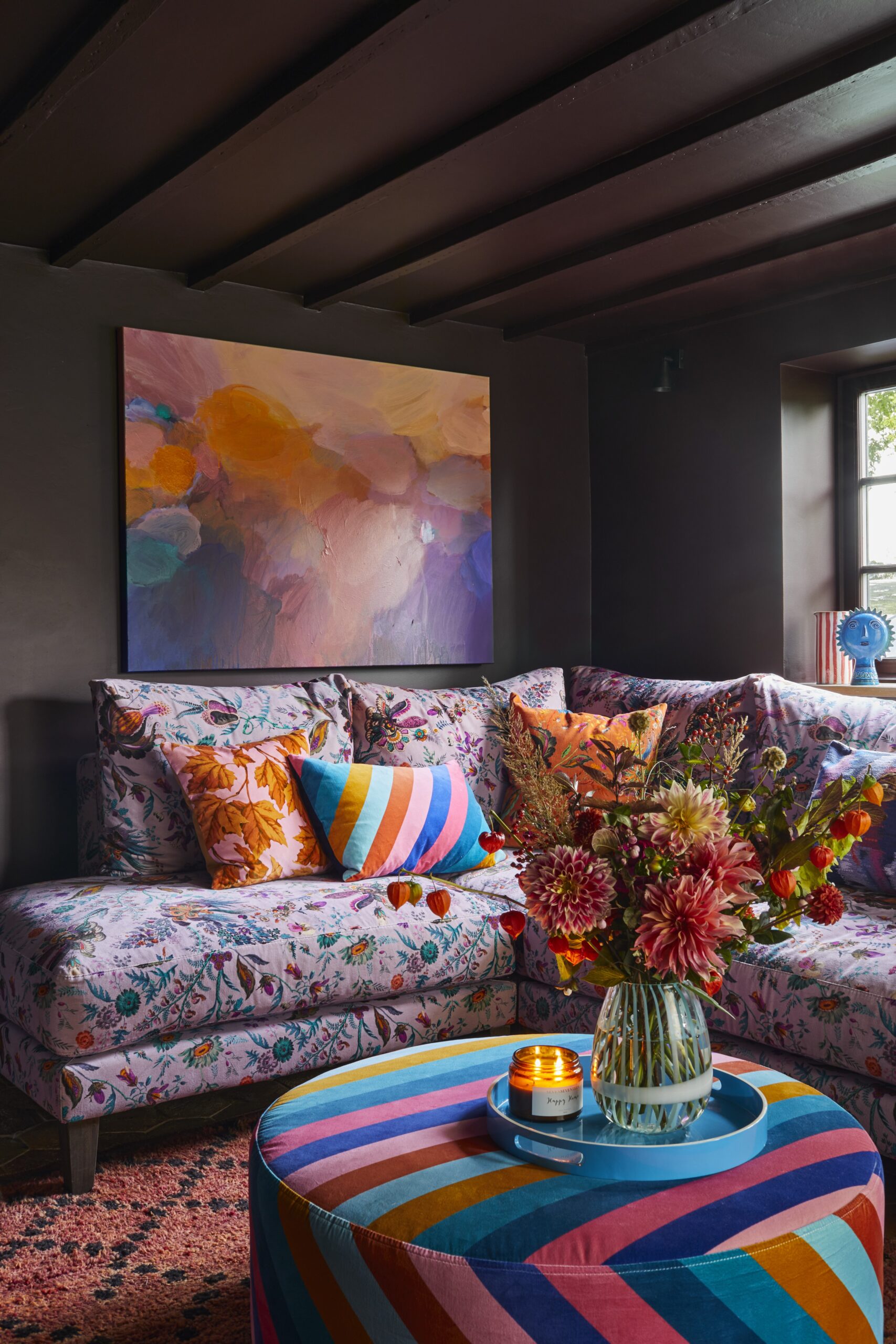 Corner of the snug with dark maroon on the walls and ceilings. Sofa in floral velvet fabric with colourful patterned scatted cushions. In front is an upholstered footstool with large floral display in a vase. 