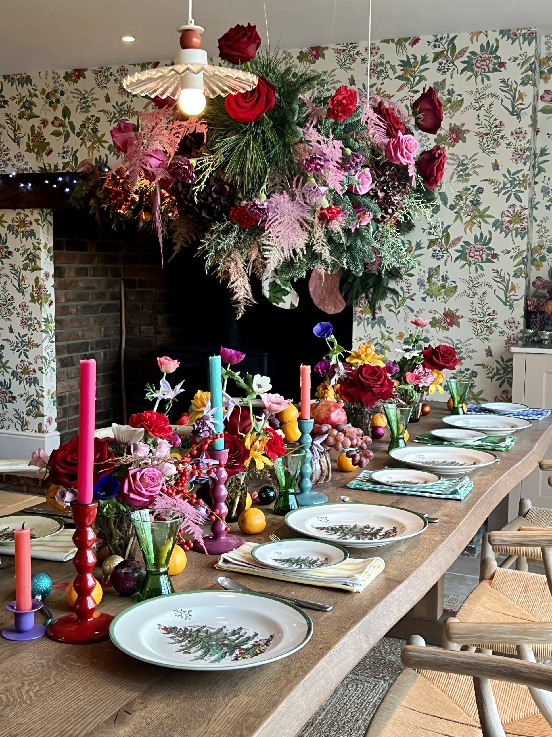 Colourful Christmas tablescape on large wooden table with Spode dinnerware which is white with a green rim and Christmas tree in the centre. Above the table hangs an extravagant foliage cloud filled with greenery, red roses and pink flowers. The centre of the table is fulled with mini floral displays. bright and colourful candlesticks with fruit and baubles scattered throughout.