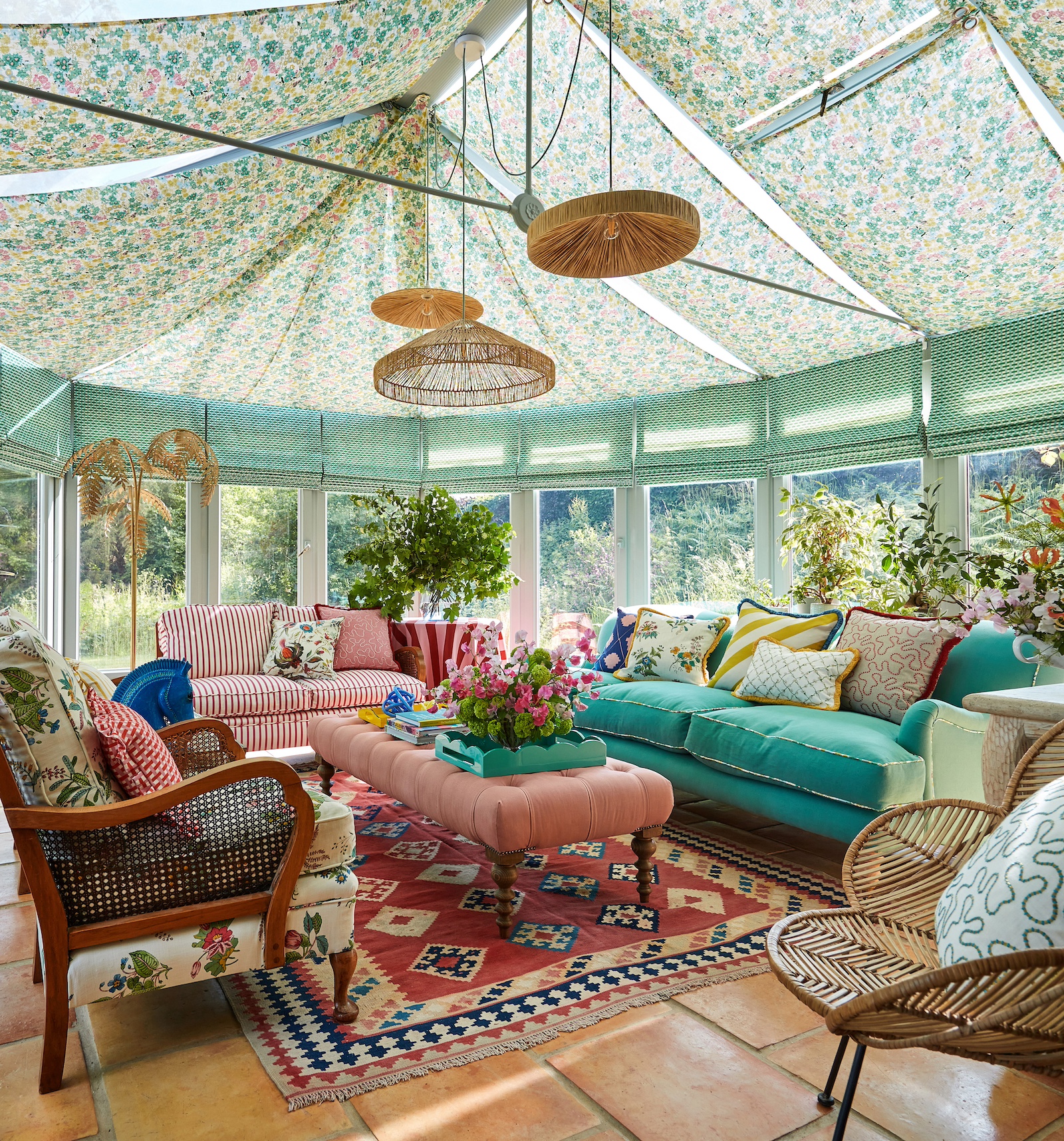 Large conservatory filled with colour sofas, armchairs, cushions and rugs. On the windows are green patterned roman blinds, half down to reveal some sunlight and on the ceiling floral fabric is draped across the glass roof to create a cosy atmosphere.  