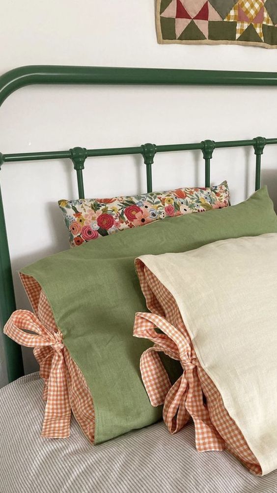 A wrought iron bed frame in green with a floral cushion and two pillows propped against it. The pillow covers are beautifully hadnmade in a green and oatmeal linen with terractorra gingham lining and bows to attach them.