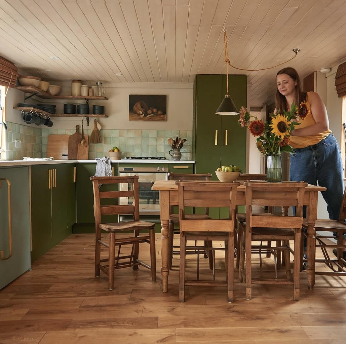 Smoky green cabinets with open shelving, green tiling and wood floor. In front of the cupboards sit an old wooden table with vintage church chairs. To the side is a white woman arranging some sun flowers in a vase.