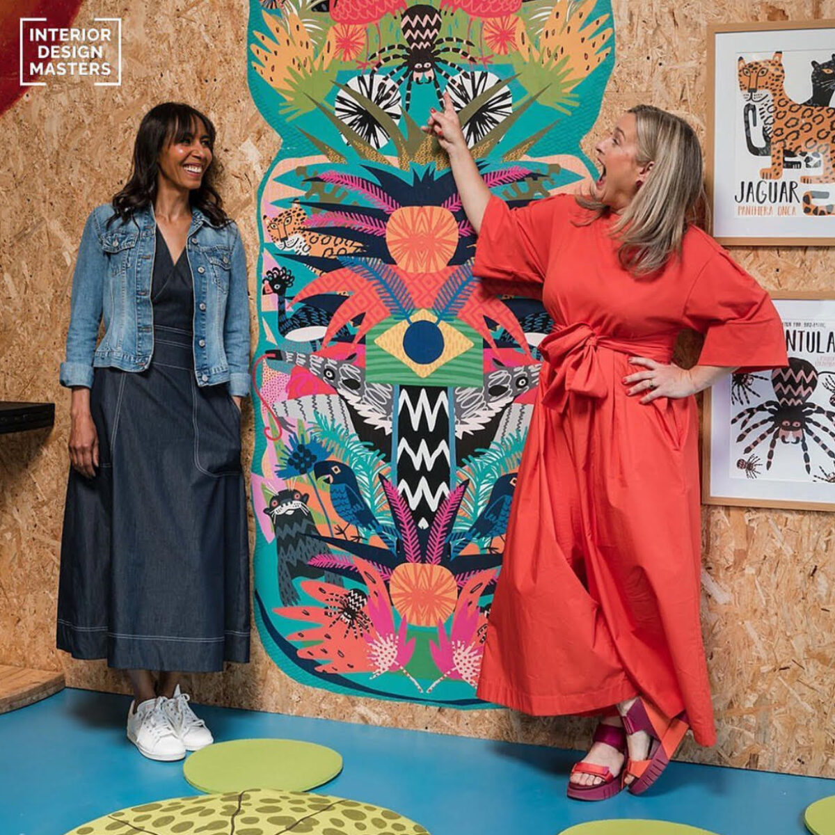 Michelle wearing navy dress and denim jacket stands next to Sophie in orange dress and heels in front of a brightly coloured display on cork walls.